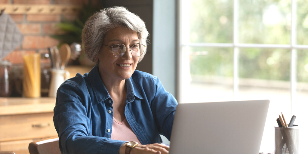 Ena has white hair and wears glasses and a jean shirt. She is sitting at her kitchen table in front of her laptop, smiling.