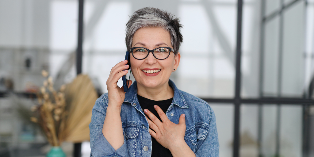 Tracy is a middle-aged woman with short grey hair and glasses. She wears a jean jacket and is holding a phone to her ear while smiling as if she just received good news.