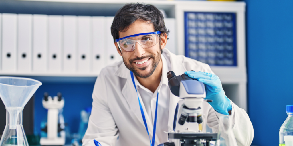 Emmanuel is a young man with dark hair and a beard. He is in a laboratory, wearing a lab coat, safety glasses and gloves. He is smiling into the camera while holding something under a microscope and taking notes.