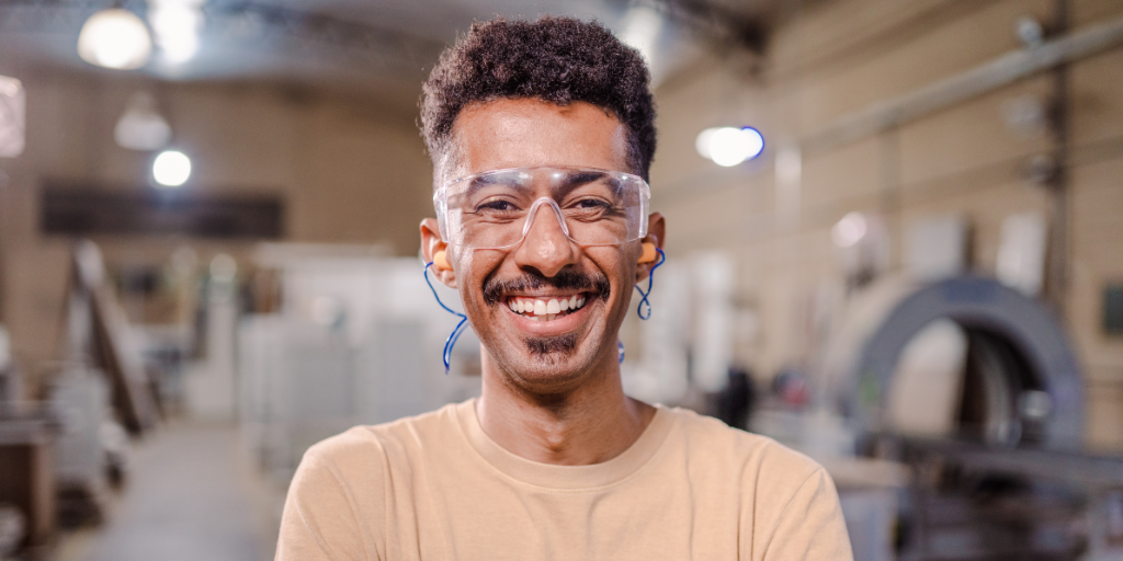 Nathan is a man in his 30s with tan skin, curly hair and a goatee. He is in a factory, wearing safety glasses and ear plugs while smiling widely towards the camera.
