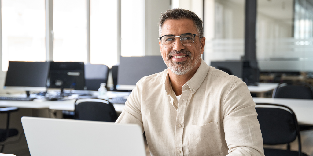 Nicholas is a middle-aged man with tanned skin, short salt and pepper hair and beard. He wears glasses and a casual shirt while sitting in front of a laptop smiling towards the camera.