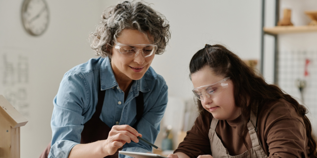 Maxine is a mature woman with short curly hair and a warm smile. She wears safety glasses and is showing a trainee with down syndrome something on am electronic tablet.