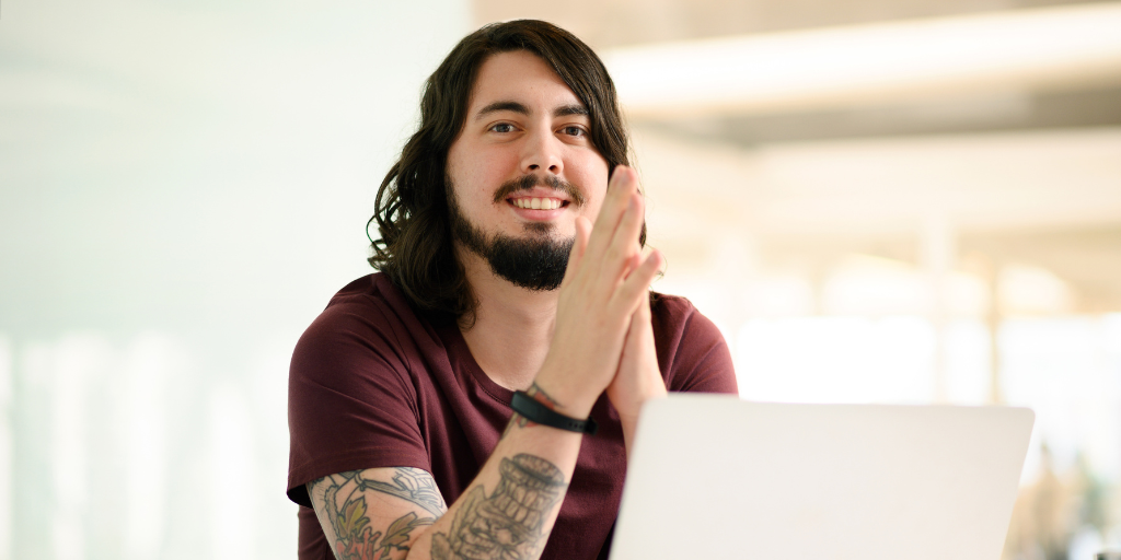 Jake is a man in his late twenties with thick shoulder-length hair and a beard. He is wearing a T shirt, has tattoos running down the length of his arm and is smiling warmly towards the camera. He has a work laptop sitting in front of him.
