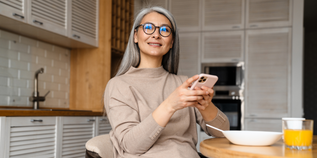 Eileen is a mature woman sitting her kitchen table. She has long straight hair and and glasses and is holding a smartphone while smiling.